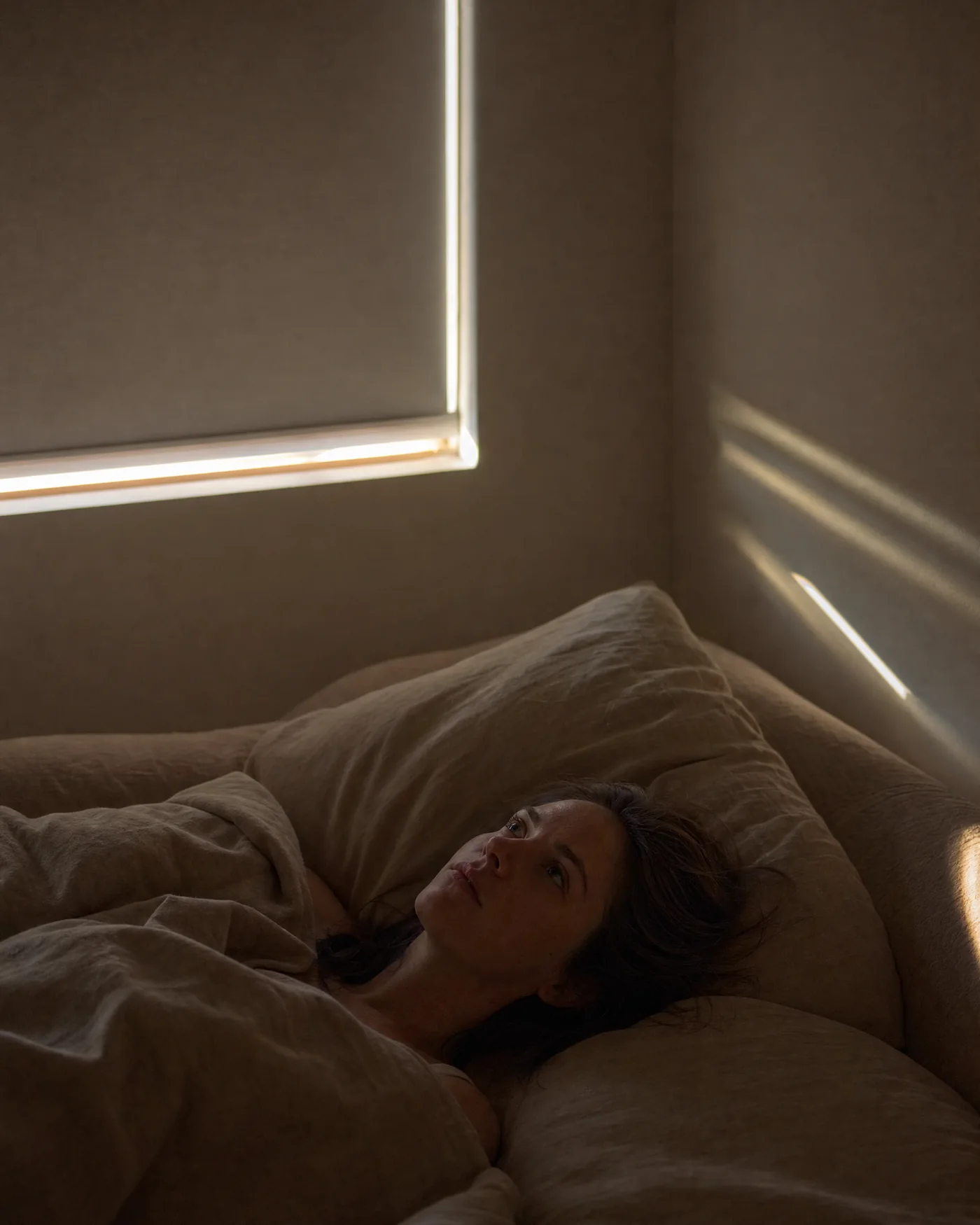 Una mujer despierta en la cama al amanecer, con la luz entrando por los bordes de la persiana